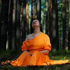 Kobieta w pomarańczowej sukni siedzi na trawie w lesie, ciesząc się chwilą kontaktu z naturą.