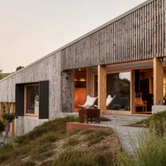 Moderner Garten am Hang mit Holzhaus, Terrasse und Sitzgelegenheiten, umgeben von üppiger Natur.