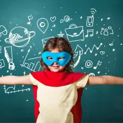Dziecko w masce superbohatera z rozłożonymi rękami na tle tablicy z rysunkami.