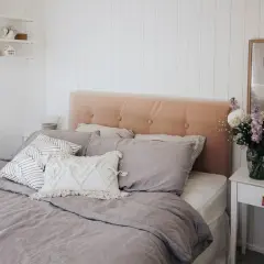 A cozy bedroom featuring a DIY upholstered headboard in a soft peach velvet, adorned with tufted buttons. The bed is dressed in linen bedding and an assortment of decorative pillows.