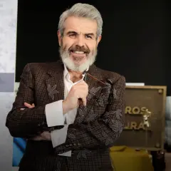 Lorenzo Caprile, con su característica barba canosa, sonríe mientras sostiene unas gafas. Viste una chaqueta de tweed con bordados florales.