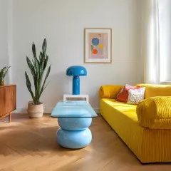 Jasny salon retro z żółtą kanapą, niebieską lampą i stolikiem kawowym. Drewniana komoda i duża roślina dopełniają przytulną atmosferę.