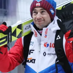 Uśmiechnięty Andrzej Stoch Jr. z nartami na ramieniu, gotowy do skoku.