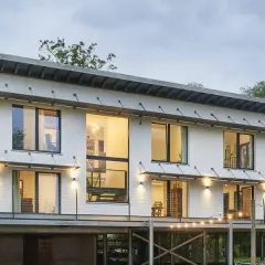 Modernes Haus auf Stelzen mit großen Fenstern und Metalltreppen, umgeben von Bäumen. Lichterketten beleuchten die Terrasse.