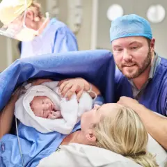 Un padre guarda con amore il suo neonato, avvolto in un asciugamano bianco, dopo un cesareo dolce. La madre sorride, circondata da personale medico.