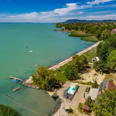 Słoneczny kemping nad Balatonem. Ludzie kąpią się w jeziorze i basenie, bawią na placu zabaw, a kampery stoją wśród drzew.