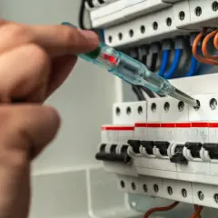 Dłoń z próbnikiem napięcia sprawdza zabezpieczenie skrzynki elektrycznej. Czerwona lampka próbnika świeci.