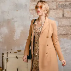 Blondynka w płaszczu w kolorze camel, idealnym dla niej. W tle stara walizka i ceglana ściana.