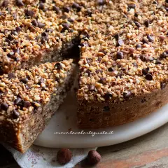 Una deliziosa torta di nocciole, ricoperta di granella di nocciole e gocce di cioccolato, con una fetta tagliata.