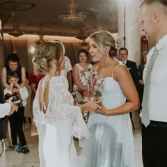 Nietypowe podziękowania dla rodziców na weselu: panna młoda w koronkowej sukni wręcza bukiet w szkle, a pan młody prezent z wstążką.