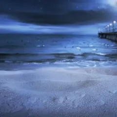 Nocna, świecąca plaża z molo i latarniami w oddali. Piasek lśni w księżycowym blasku.