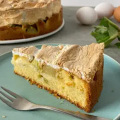 Ein saftiges Stück Rhabarber Baiser Kuchen mit einer luftigen Baiserhaube liegt auf einem blauen Teller. Im Hintergrund ein ganzer Kuchen und frischer Rhabarber.