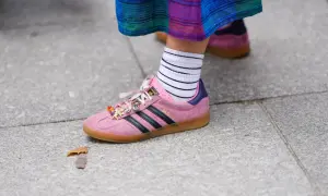 Różowe buty sportowe Adidas Gazelle z czarnymi paskami i charmsami, białe skarpetki w paski. W tle widać drugie, podobne buty w innym kolorze.