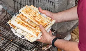 Talerz z grillowanymi wrapami kebab, trzymany przez ręce na tle rusztu i kamieni.