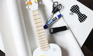 Przygotowania do wysyłki gitary: biała gitara, nożyczki, klej, zszywacz i ozdobna kokardka.