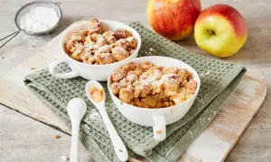 Zwei Schalen mit warmem Apfel Crumble, bestäubt mit Puderzucker. Daneben liegen zwei Äpfel und Löffel.