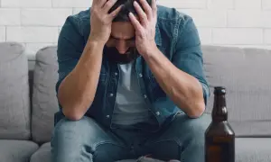 Man met handen in het haar, zittend op een bank met een fles bier. Dit illustreert de definitie van verslaving: een dwangmatige behoefte.