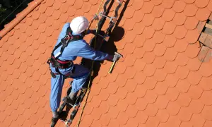 Pracownik w niebieskim kombinezonie i białym kasku wykonuje mocowanie liny na dachu. Używa młotka i zabezpieczeń.