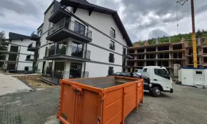 Pomarańczowy kontener na gruz przed nowym budynkiem i placem budowy.