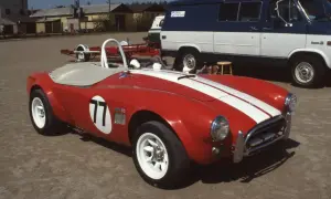 Röd sportbil, en kungens AC Cobra med nummer 77, redo för race. Bakom skymtar en blåvit skåpbil.