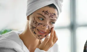 Femme souriante appliquant un gommage sur son visage. Elle porte une serviette sur la tête et un peignoir. La question est : combien de temps laisser un gommage sur le corps ?