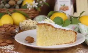 Una fetta di soffice torta caprese al limone, guarnita con zucchero a velo e foglioline di menta, circondata da limoni freschi e mandorle.