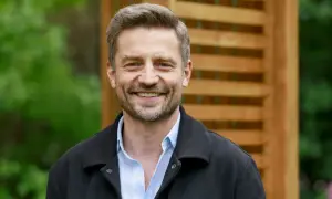 Uśmiechnięty Marcin Rogacewicz, znany z wielu filmów, pozuje na tle drewnianej pergoli i zieleni.