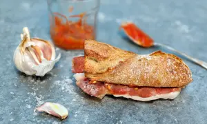 A delicious bocadillo de jamon, with a crusty baguette filled with cured ham, sits on a slate surface next to garlic and a spoonful of tomato spread.
