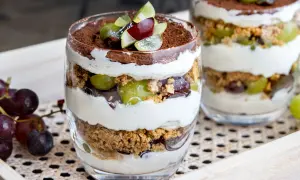 Dessert im Glas mit Schichten aus Creme, Keksbröseln, grünen und roten Trauben, garniert mit Kakaopulver.