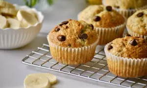 Frisch gebackene schoko bananen muffins mit Schokostückchen auf einem Kuchengitter. Daneben eine Schüssel mit Bananenscheiben.