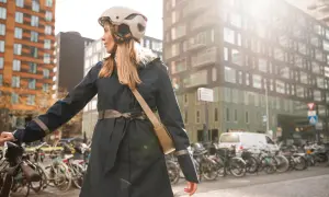 Kobieta w stylowej, granatowej kurtce przeciwdeszczowej z odblaskowymi elementami, w kasku rowerowym, stoi obok roweru na tle miejskiej zabudowy.