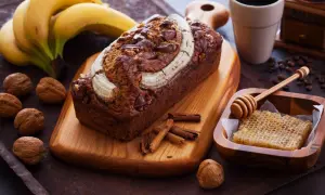 Ein saftiges Bananenbrot mit Schokoladenstückchen und Nüssen, garniert mit einer Banane. Daneben Kaffee, Honigwabe und Zimtstangen – ein Genuss, der auch bananenbrot gesund macht.