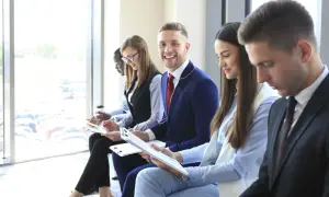 Giovani professionisti in attesa per un colloquio di gruppo, alcuni con appunti, uno sorride.