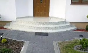 Nowe schody z żywicy na balkon, z kamyczkami, prowadzące do drewnianych drzwi. Wokół kostka brukowa i roślinność.