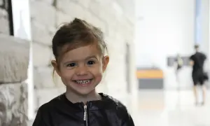 Uśmiechnięta dziewczynka z zespołem Williamsa w czarnej kurtce, z jasnymi włosami i dużymi oczami, na tle jasnej ściany i rozmazanych postaci.