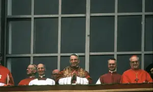 Kardynałowie w czerwonych szatach i jeden w białej sutannie, z papieżem w centrum. W tle kratowane okno.