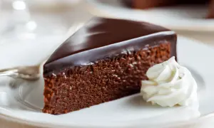Ein Stück Sachertorte, wie im Sachertorte Rezept Original Lafer, mit glänzender Schokoladenglasur und einem Klecks Schlagobers auf einem weißen Teller.