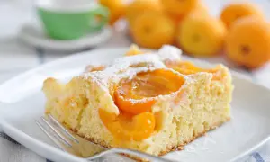 Una fetta di torta albicocche e yogurt, soffice e profumata, cosparsa di zucchero a velo. Sullo sfondo, albicocche fresche e una tazzina verde.
