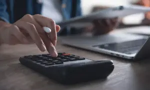 Dłoń wpisuje dane na kalkulatorze, by obliczyć, ile wynosi trzynastka. Obok leży laptop i tablet.