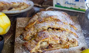Strudel di pasta frolla dorato, cosparso di zucchero a velo, con ripieno di mele e uvetta. Accanto, scorza di limone e una tazza di uvetta.