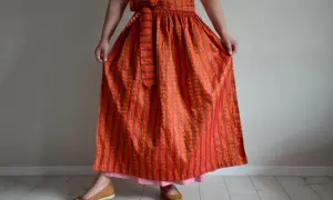 Eine Frau trägt ein Dirndl mit einer leuchtend orangefarbenen Dirndlschürze, die mit Streifen und kleinen Tupfen gemustert ist.