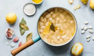 Top-down Ansicht eines Topfes mit weißen Riesenbohnen in Brühe, daneben Zitronen, Knoblauch und Lorbeerblätter.