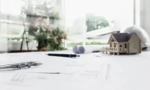 Model domu, cyrkiel, ołówek i plany architektoniczne na biurku. W tle widać zieleń i budynki, sugerujące drugą linię zabudowy.