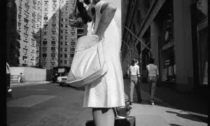 Una donna con borsa a tracolla cammina per strada, con palazzi alti sullo sfondo. Mark Cohen photography cattura la vita urbana.