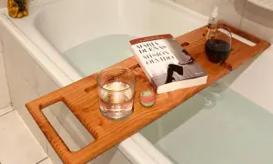 Tabla de madera sobre bañera con libro, vaso de agua, bomba de baño y copa de vino. Preparando un baño relajante.