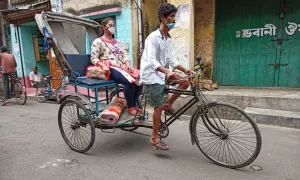Rowerzysta w masce wiezie kobietę w rikszy. Typowy transport w Indiach, uliczny krajobraz z psami i budynkami.