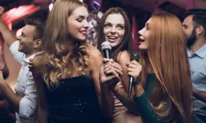 Trzy kobiety śpiewają do mikrofonów, bawiąc się przy karaoke. W tle widać innych ludzi tańczących.
