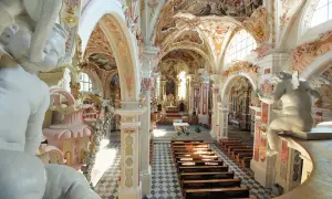 Prunkvolle Deckenmalereien und Stuckaturen im Kloster Neustift. Engelstatue blickt auf Kirchenbänke und den Altar.