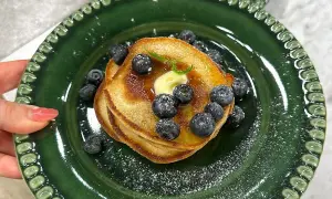 En hög med amerikanska pannkakor toppas med smör, blåbär och sirap på en grön tallrik.