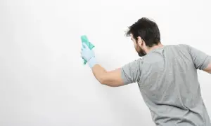 Un homme en gant bleu nettoie un mur blanc avec un chiffon vert. Il montre comment nettoyer un mur blanc sans enlever la peinture.